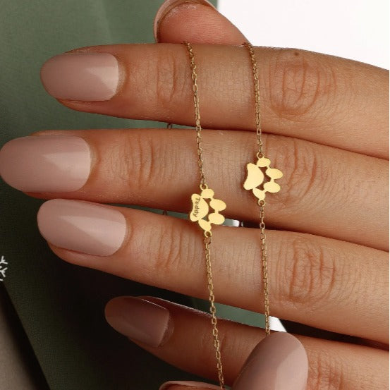 Gold paw-print bracelet engraved with a pet's name, worn on a hand next to a plain gold paw-print bracelet